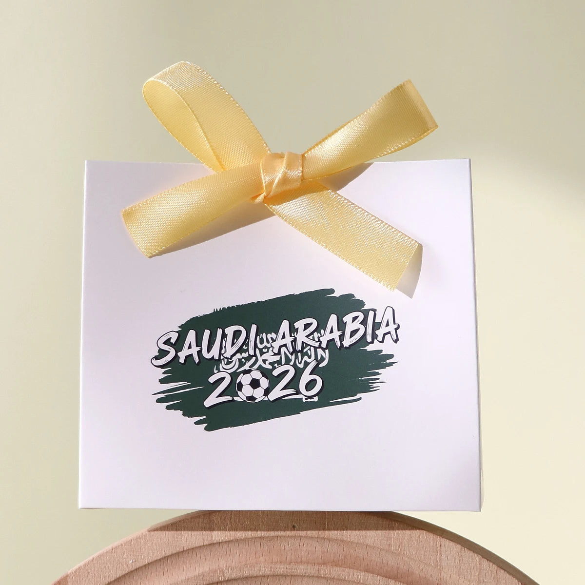 Saudi Arabia 2026 Football-Themed Gift Box, Simple White Box with Green Football Print, Adorned with a Delicate Yellow Satin Ribbon Bow, Featuring a Clean and Elegant Design The Box Has Clear Printing and a Fine Texture, Maki Qme