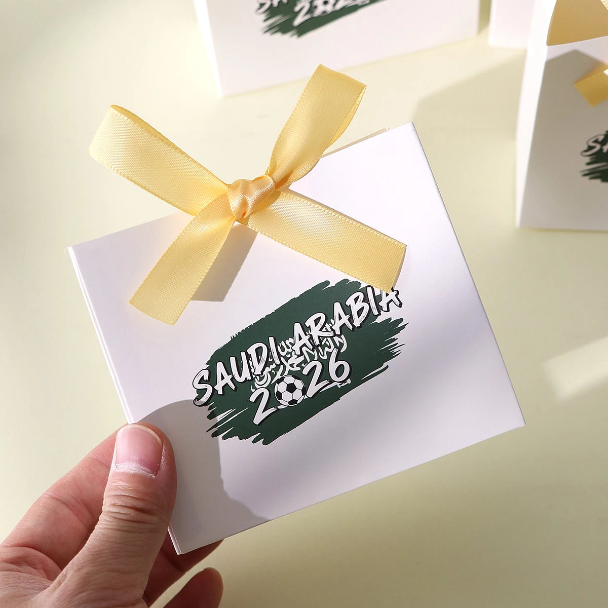 Saudi Arabia 2026 Football-Themed Gift Box, Simple White Box with Green Football Print, Adorned with a Delicate Yellow Satin Ribbon Bow, Featuring a Clean and Elegant Design The Box Has Clear Printing and a Fine Texture, Maki Qme